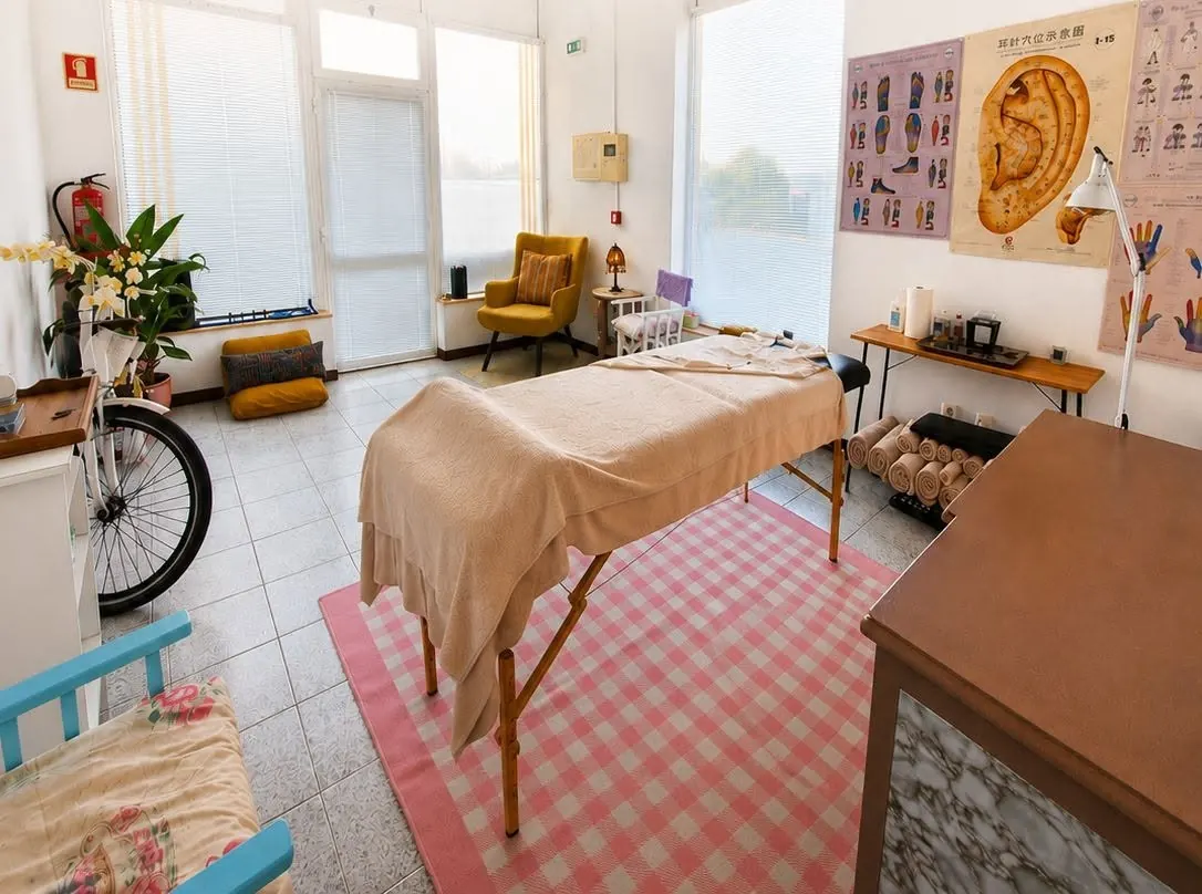 Rainbow Tree therapy room in Vila Nova de Poiares: massage table, pink gingham rug, yellow armchair, plants and reflexology and auriculotherapy charts on the wall